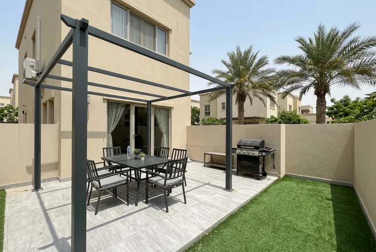 A clean Dubai villa garden terrace with a pergola, cushioned outdoor dining chairs and artificial grass in cool-season light